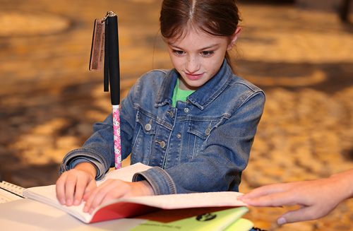 A young blind girl with a white cane reads a Braille book.